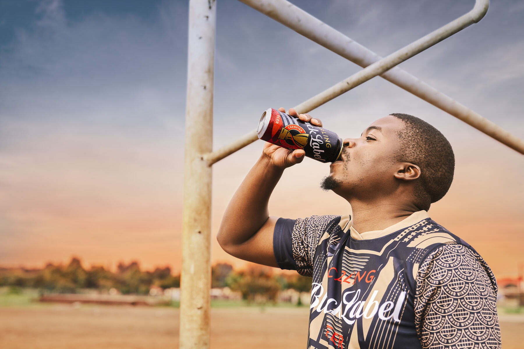 Man drinking a Carling Black Label at sunset beside football goalposts