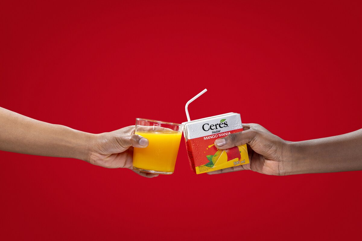 Two hands toasting with a glass of juice and a Ceres Nectar Mango Mania carton