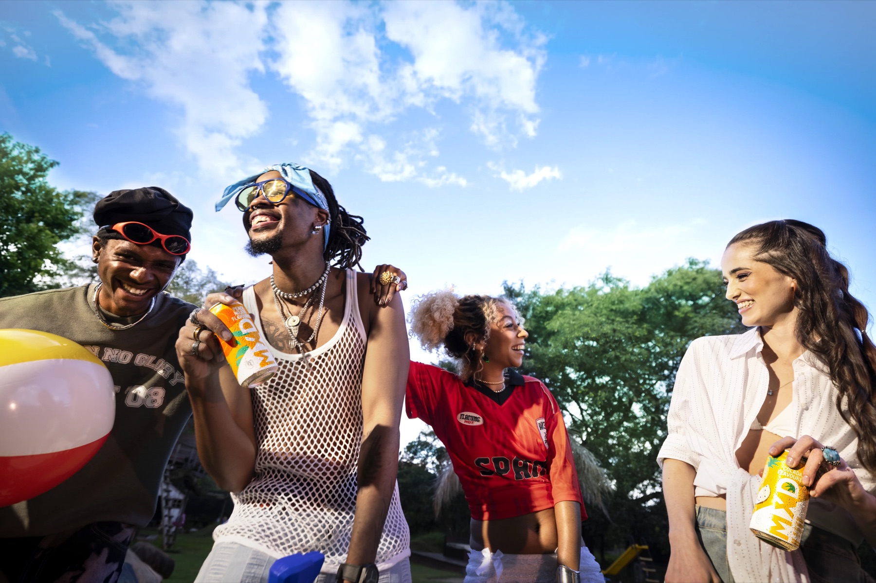 Four friends laughing with MXD spirit cooler cans and a beach ball under a summer sky