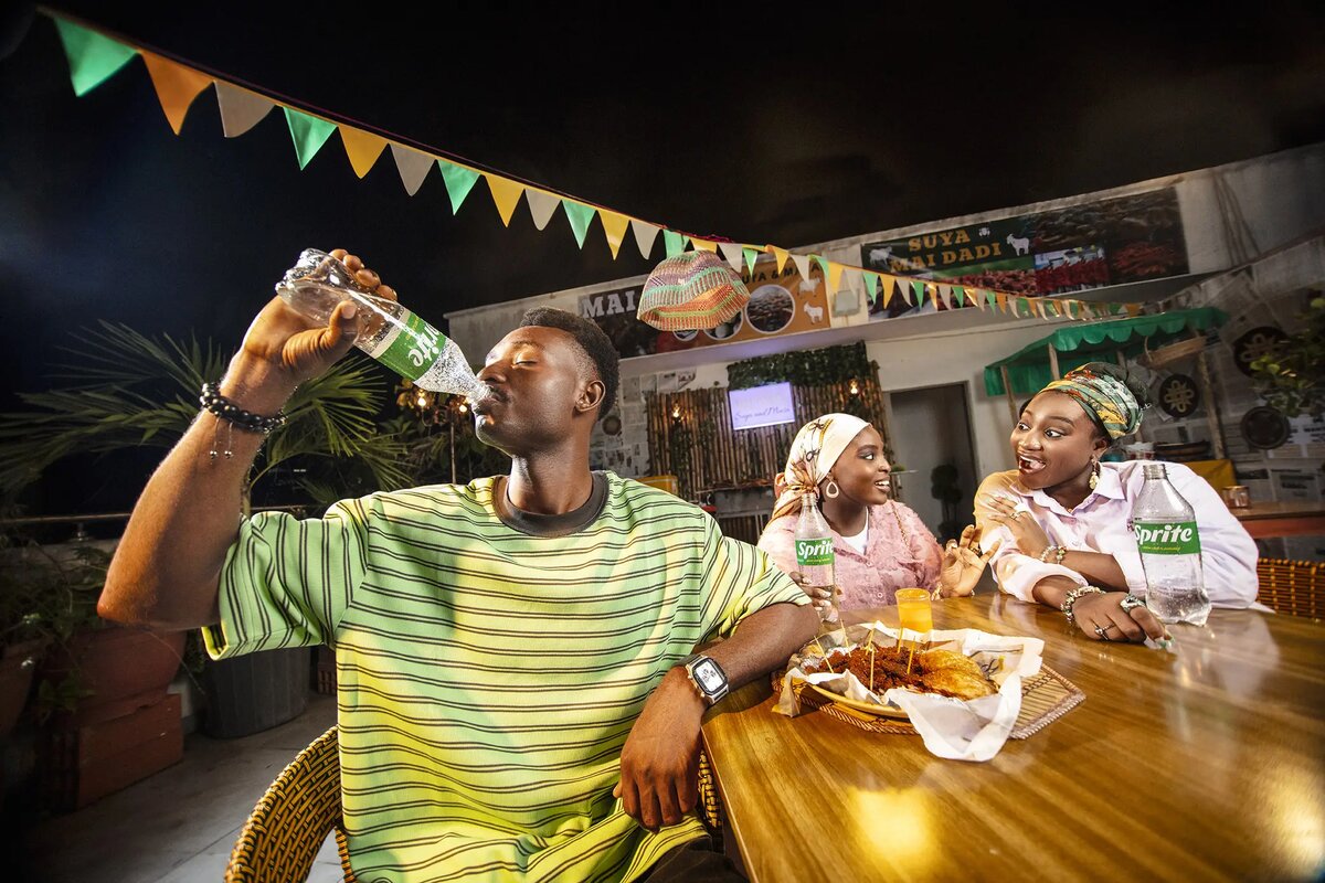 Friends drinking Sprite at a suya food stand under festival bunting at night