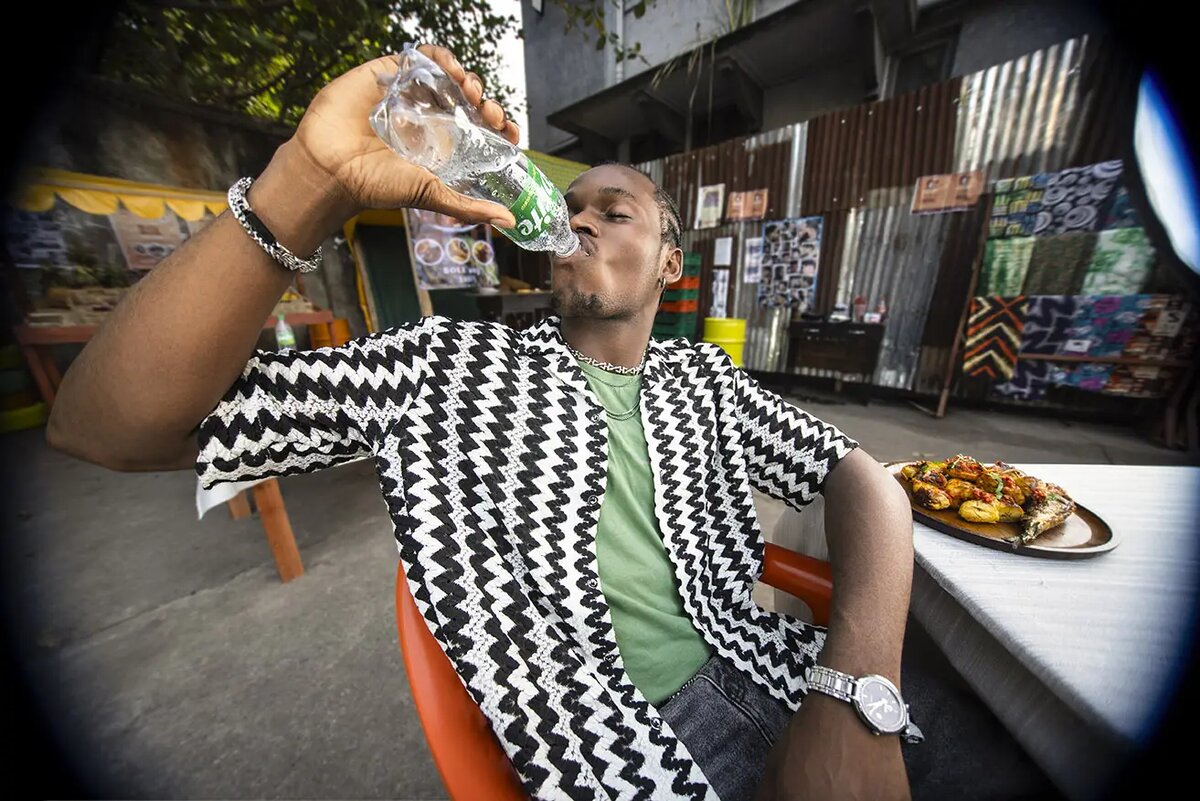 Fisheye point-of-view shot of a man drinking Sprite beside a suya platter